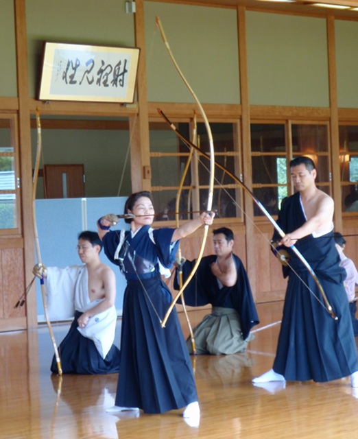 A woman dressed in a hakama stands with bow drawn, while other archers are kneeling down in the background