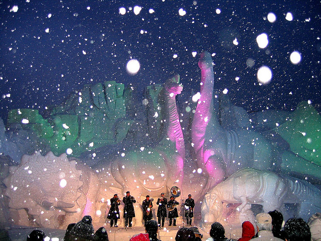 A band performing on a stage in front of a giant dinosaur sculpture during a snowstorm
